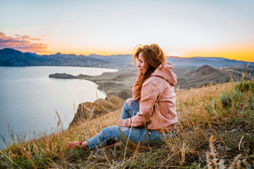 Fototapeta premium A beautiful young woman tourist is sitting on top of a mountain and enjoying the view of the sunset sea and mountains. The concept of freedom and meditation, an active lifestyle.