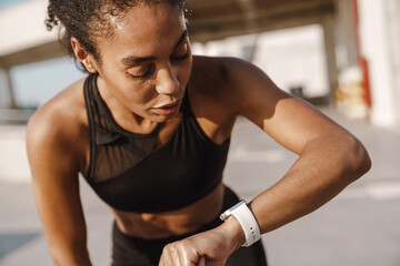 Fototapeta na wymiar Black sportswoman looking at smartwatch while working out on parking
