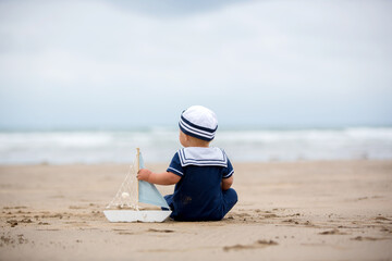 Fototapeta na wymiar Baby boy sitting on the beach near the water and plays with a toy ship and bear toy