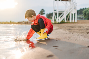 Boy in a red raincoat and yellow rubber boots playing with water at the beach. School kid in a...