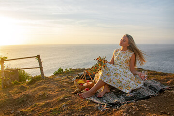 A young woman is resting by the sea on a picnic, drinking water from a glass.