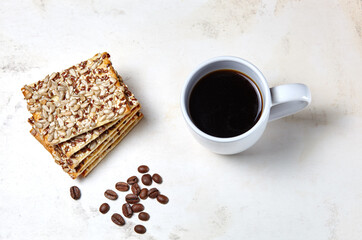 White cup with coffee, coffee beans and crunchy crispbread on a wooden background, closeup. Tasty breakfast, morning routine concept, top view