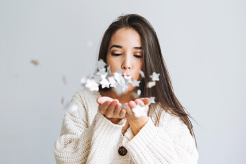 Young asian woman in white knitted cardigan blows on snowflakes in her hands on grey background, winter christmas time