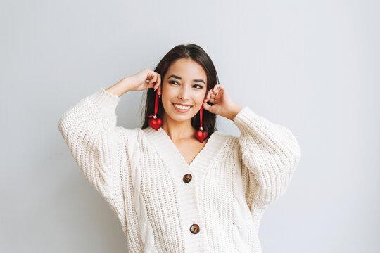 Young happy asian girl with long hair in white knitted cardigan with red heart christmas ball in her hands on grey background, winter christmas time