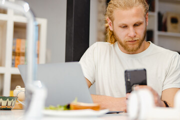 Bearded blonde man using cellphone and laptop while sitting on table