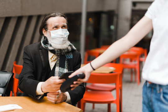 White senior man in face mask paying with mobile phone at cafe