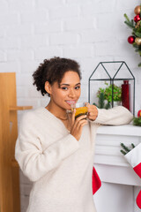 Smiling african american woman holding cup of tea near fireplace with christmas stockings