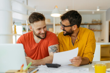 A gay couple calculating their monthly income. There is so much to pay, taxes, bills. They are looking at bills and smiling. - 462440438