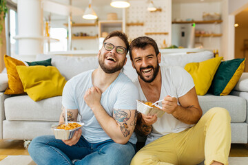 Happy hipster gay married couple sitting on the floor at home, eating pasta and watching tv...