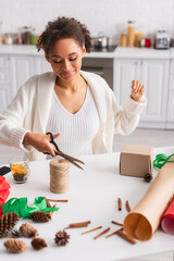 Young african american woman cutting twine while decorating gift near tea and cinnamon sticks