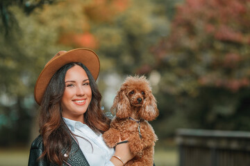 A beautiful young dark-haired woman walks in the park and hugs her poodle dog. Autumn mood.