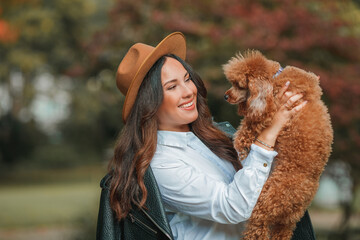 A beautiful young dark-haired woman walks in the park and hugs her poodle dog. Autumn mood.