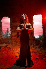 Fototapeta premium A brunette girl with long hair in an evening dress in a vintage Gothic castle. A woman who looks like an evil witch or sorceress in an abandoned old church. Female Model posing in Halloween