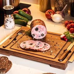 Smoked ham stuffed with black olives on a wooden board. Fresh vegetables and bread on the kitchen table around