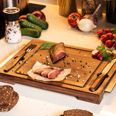 Smoked ham on a wooden board among vegetables and bread on the kitchen table. Fresh breakfast ingredients