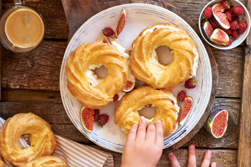 Choux rings with cream, figs and raspberries. Wooden background, top view. Children's hand holding a choux ring.