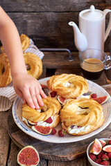 Choux rings with cream, figs and raspberries. Wooden background, side view. Children's hand holding a choux ring.