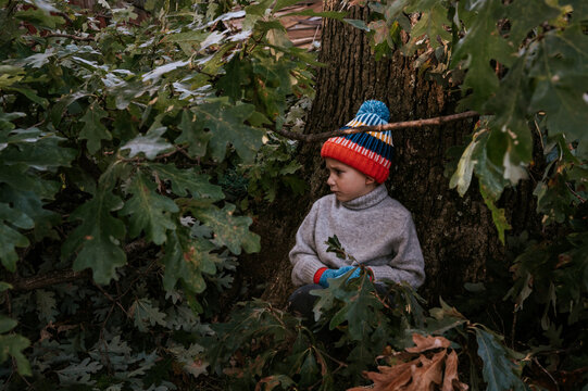 Sad little boy in hat and mittens sitting under a tree