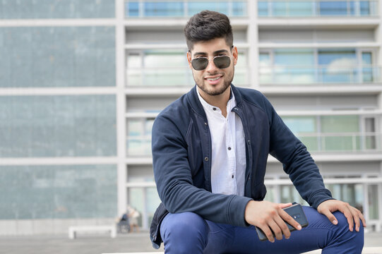 Portrait of young man in stylish sunglasses posing while sitting in bench in urban background. High quality photo