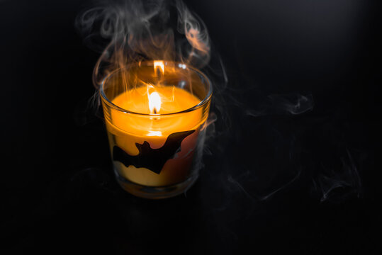 Halloween mystical decor with a lit candle in a glass beaker depicting a bat on a black background. Halloween holiday mystical smoke from a candle.