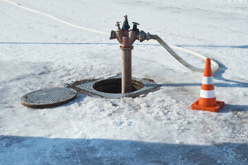 A shabby fire hydrant in an open hatch and a traffic cone nearby, in the middle of a snowy winter pavement.