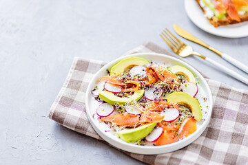 Flat lay top view of healthy salad with smoked salmon, avocado, radish and fresh microgreens alfalfa sprouts on white plate on gray concrete background. Healthy lifestyle. Growing sprouts. Green