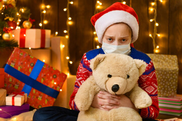 Fototapeta na wymiar Child girl as Santa helper posing in new year decoration. Wearing a protective face mask against viruses, coronaviruses. She's playing with a toy bear. Holiday lights and lots of gifts