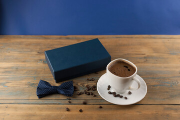A cup of coffee with bow tie and gift box on old wooden table with some beans. Father day card.