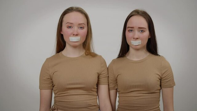 Portrait of twin sisters with taped mouth looking at camera with sad facial expression. Identical Caucasian young slim beautiful women posing at grey background. Speechlessness and human rights