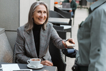 Grey white senior woman paying with mobile phone in cafe