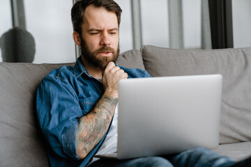 Bearded european man working with laptop while sitting on sofa at home