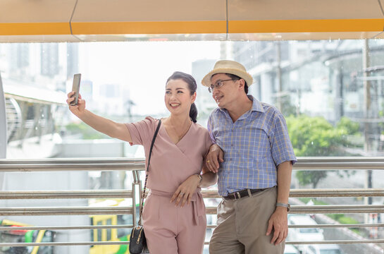 Senior couple traveling together and taking a selfie, with bag and hat on city blur background