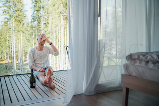 Portrait of a tired Middle-aged man sitting barefoot on forest house balcony floor,he drinking white wine and massaging temples. Exhausted people or mental health or personal problems concept image.