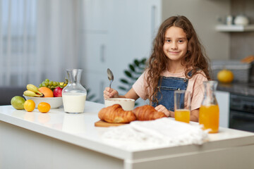 Adorable little girl, eating cereals healthy breakfast on the kitchen, looking at the camera with...