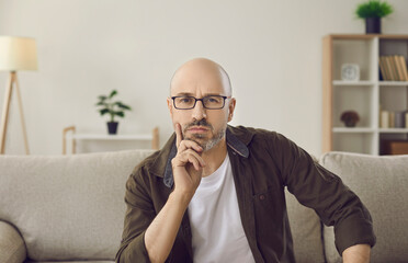 Portrait of serious pensive intelligent man on sofa at home. Bald adult man in glasses sitting on...