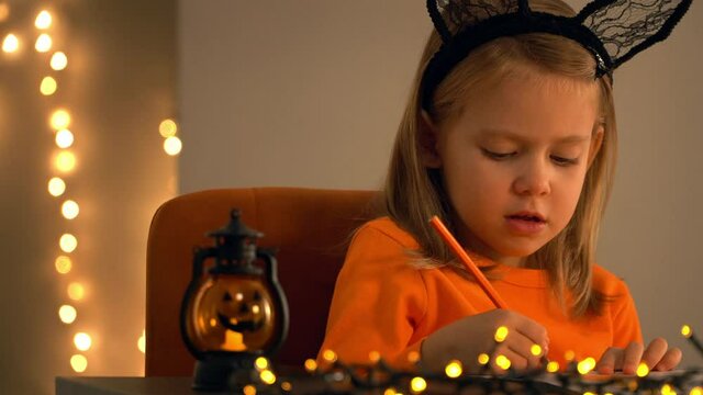 Little girl sits in dark with pumpkin lamp and makes decoration for holiday Halloween. Child paints orange pencil on background of shining blurred lights of golden garland.