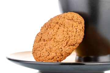 One fresh oatmeal cookie on a black ceramic saucer with a cup, close-up, isolated on white.