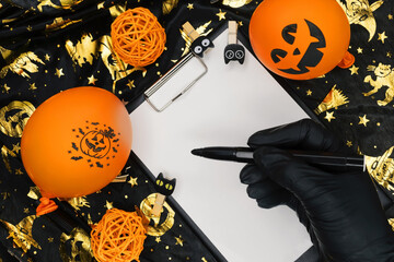 Halloween concept. A black-gloved hand holds a felt-tip pen over a white blank sheet. An orange...