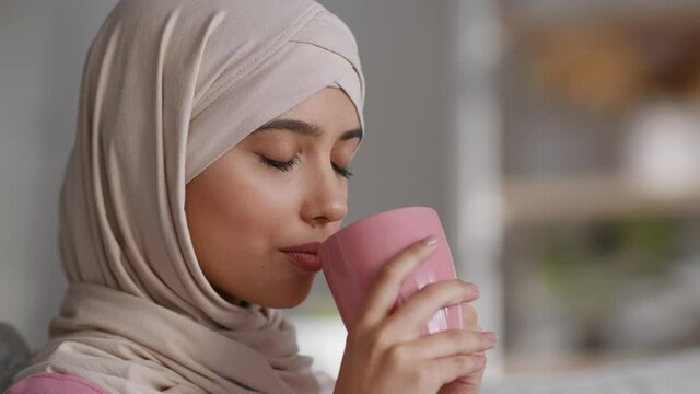 Morning relax. Close up of young peaceful muslim woman in hijab drinking hot coffee, enjoying fresh drink at home