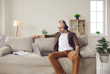 Obraz premium Serious bald bearded mature man in glasses and headphones sitting on sofa at home, using laptop computer, watching video lecture, working on business project, listening to music and staying focused