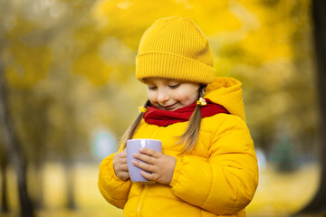 Warm fall photo. Close-up shot of a pretty cute little child girl wearing bright yellow coat and cap, holding a cup of hot chocolate or tea in autumn golden park