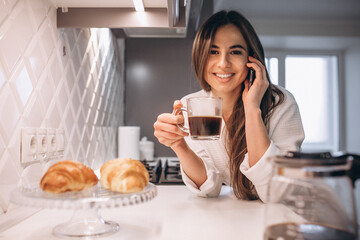 Woman drinking coffee with croissant at home and using phone