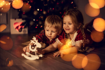children - boy and girl are playing on the floor with toy deer. Christmas tree, orange bokeh, new year lights, gifts, santa claus. Childhood, magic, anticipation of the holiday, advent calendar