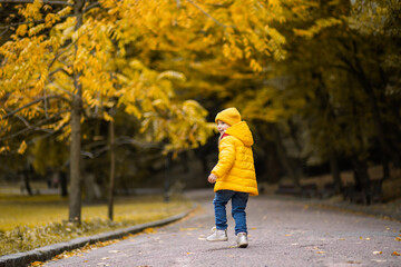 Autumn in the park. Back view of little cute girl in stylish yellow coat and cap, running, jumping and having fun, while walking in beautiful golden autumn park