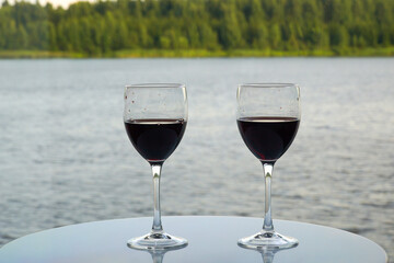 Two glasses with wine on a glass table against the background of water and forest shore.