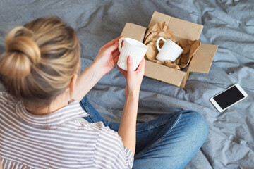 Woman sits on the bed, unpacks a cardboard box with dishes, buys goods online, delivery concept.