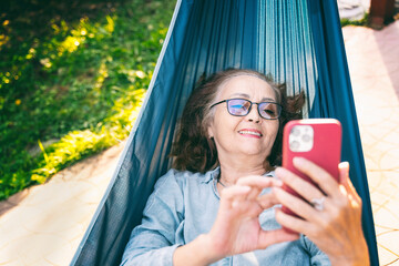 Happy beautiful senior woman smiling happy relaxing on a hammock at home using smartphone