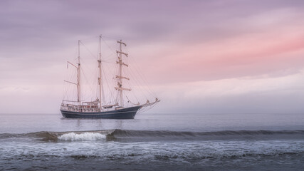 Rolling wave in front of beautiful tall ship covered in haze with soft morning sky, anchored near...