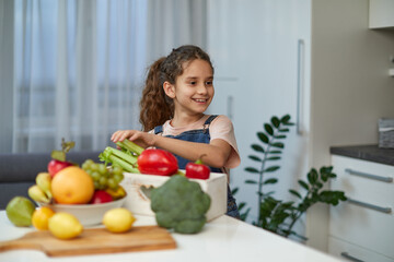 Adorable laughing and curly little girl sits at the table in the kitchen, near vegetables.