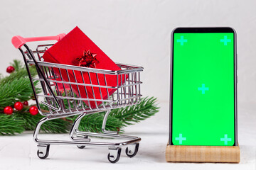 Festive background with shopping cart with red gift box, smartphone with green screen and Christmas...